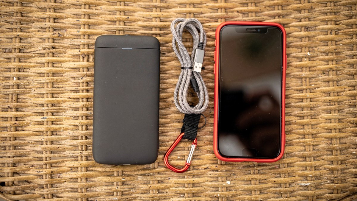 A mobile hotspot device, a smartphone with a red case, a coiled USB cable, and a red carabiner laid flat on a woven rattan cafe table.