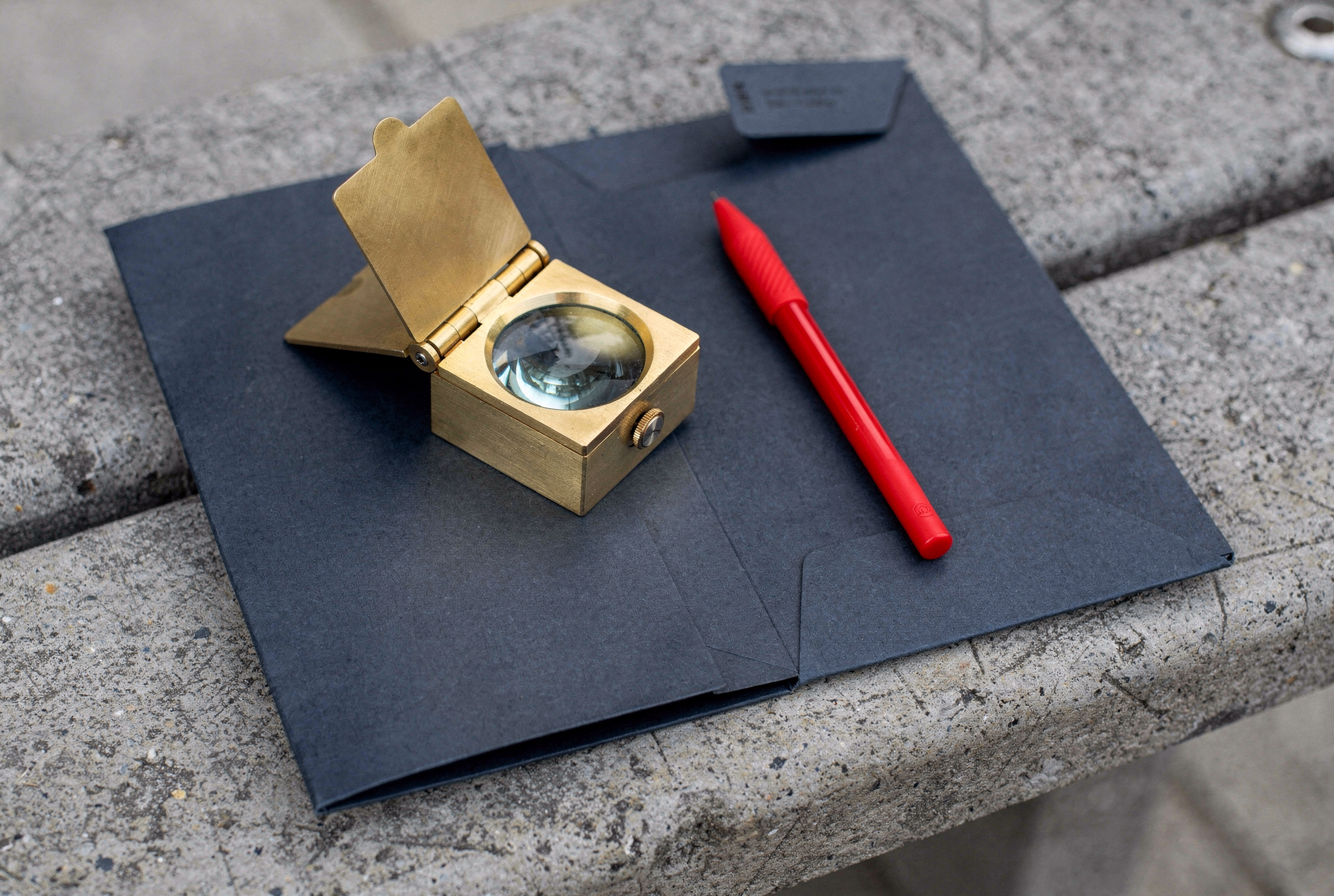 A brass pocket magnifying loupe and a red stylus pen resting on a dark blue folder over a concrete bench.