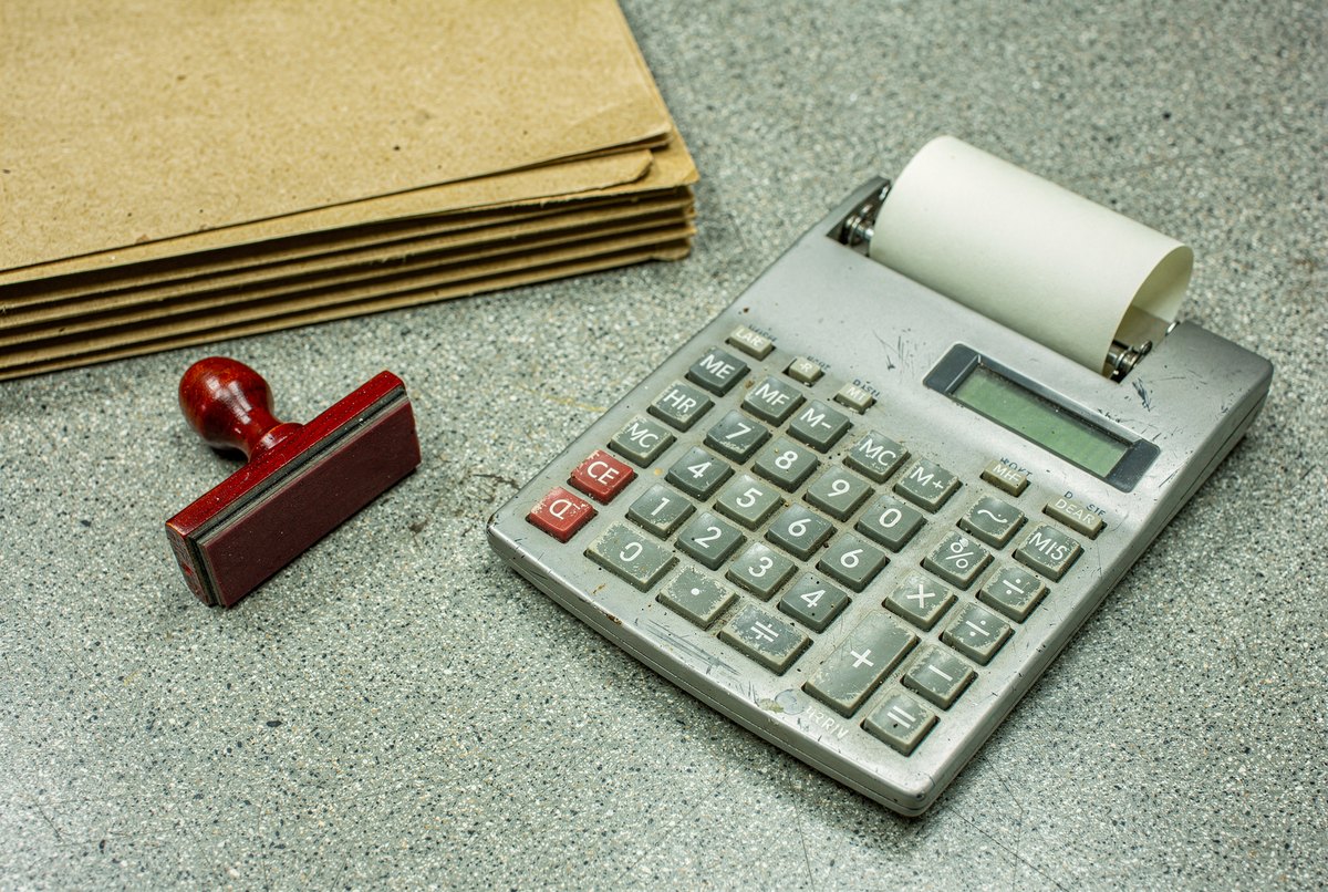 A grey desktop calculator with paper tape resting next to a red-handled stamp on a linoleum surface under cool office light.