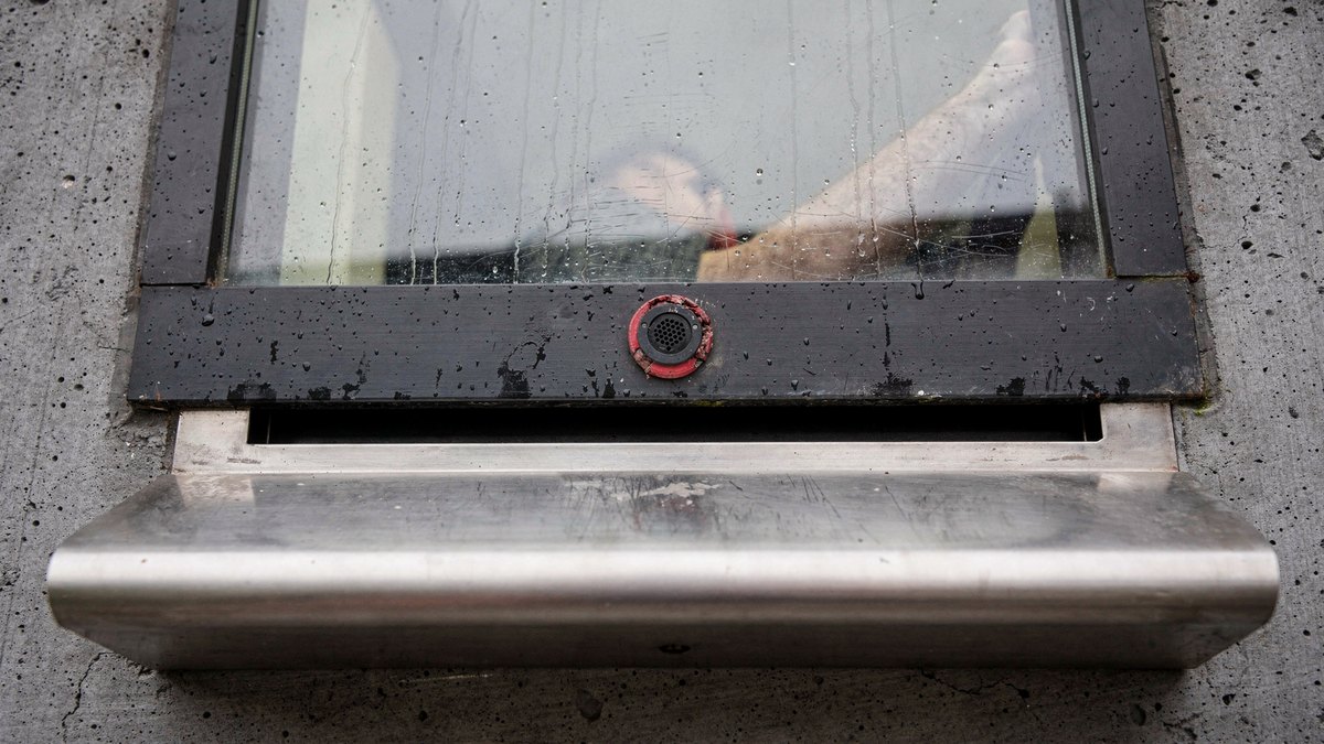 A weathered stainless steel document pass-through tray and security glass window installed in an exterior concrete wall on an overcast day.