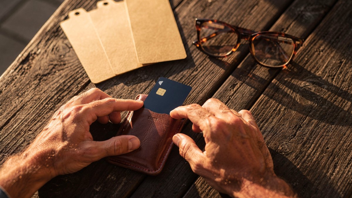 A pair of hands sliding a dark bank card into a worn leather card sleeve on a rustic wooden table during golden hour.