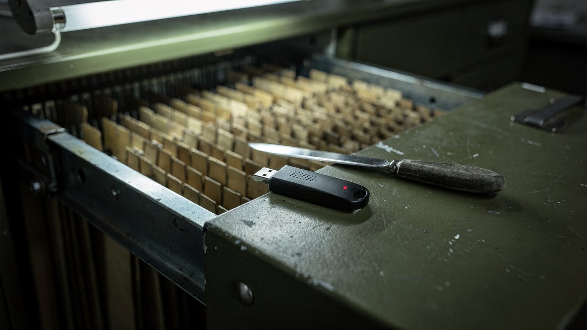 A hardware security key and stainless letter opener resting on an open metal filing drawer under green-tinted office fluorescent lighting.
