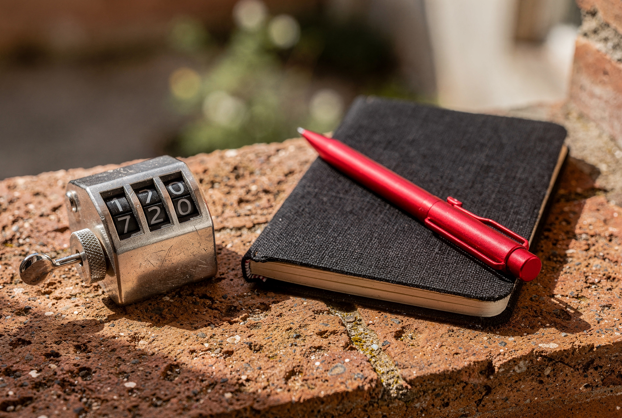 A silver mechanical tally clicker and a red metal stylus resting on a closed black linen notebook on a terracotta windowsill in morning sun.
