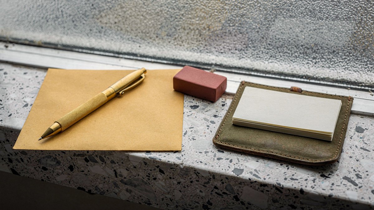 A brass drafting pencil resting on a kraft paper envelope next to a red rubber stamp on a terrazzo windowsill.