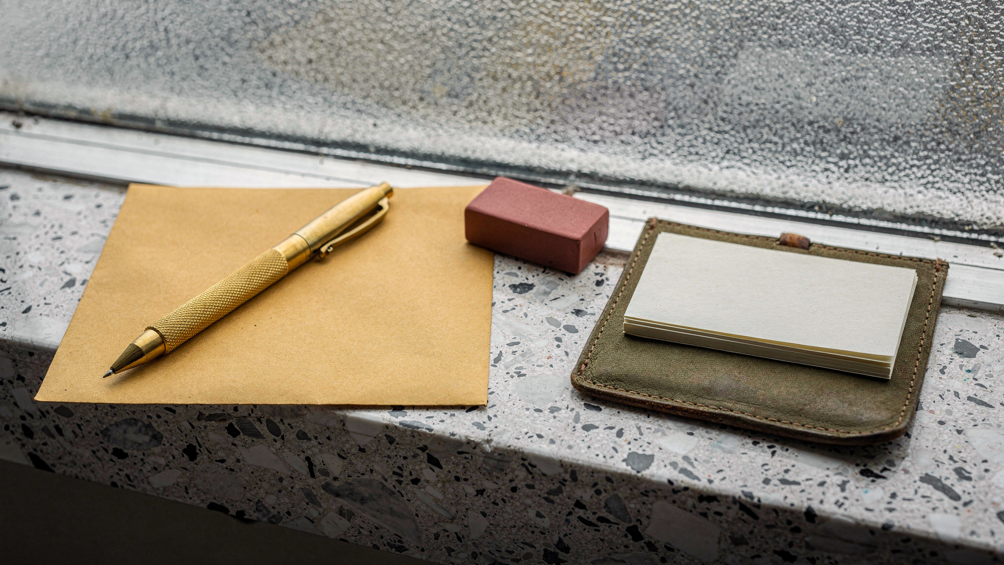 A brass drafting pencil resting on a kraft paper envelope next to a red rubber stamp on a terrazzo windowsill.