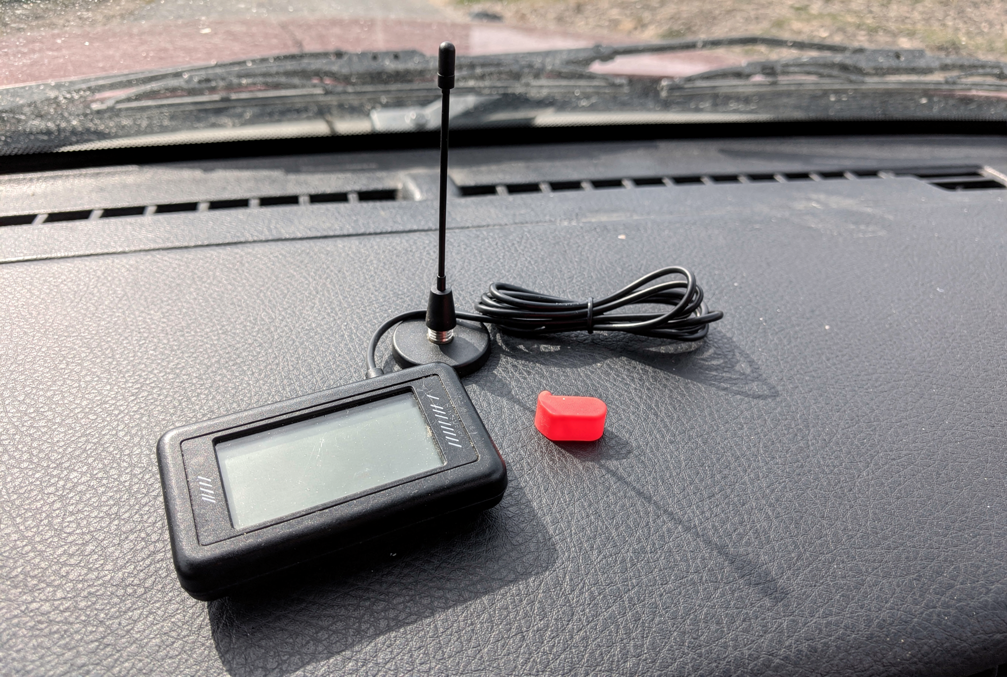 A handheld signal meter, small whip antenna, and red rubber port cap resting on a vehicle dashboard.