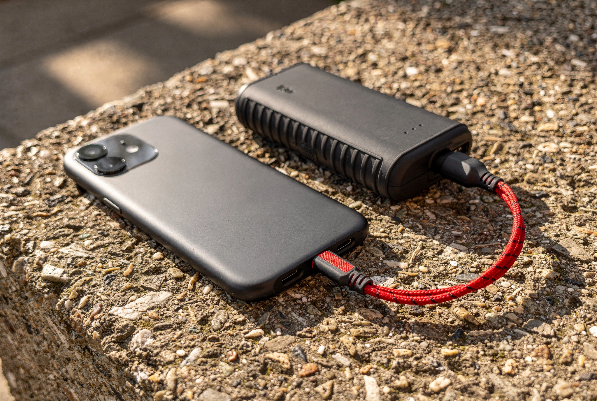 A grey smartphone and a rugged charcoal power bank connected by a short red braided cable on a concrete bench in bright morning light.