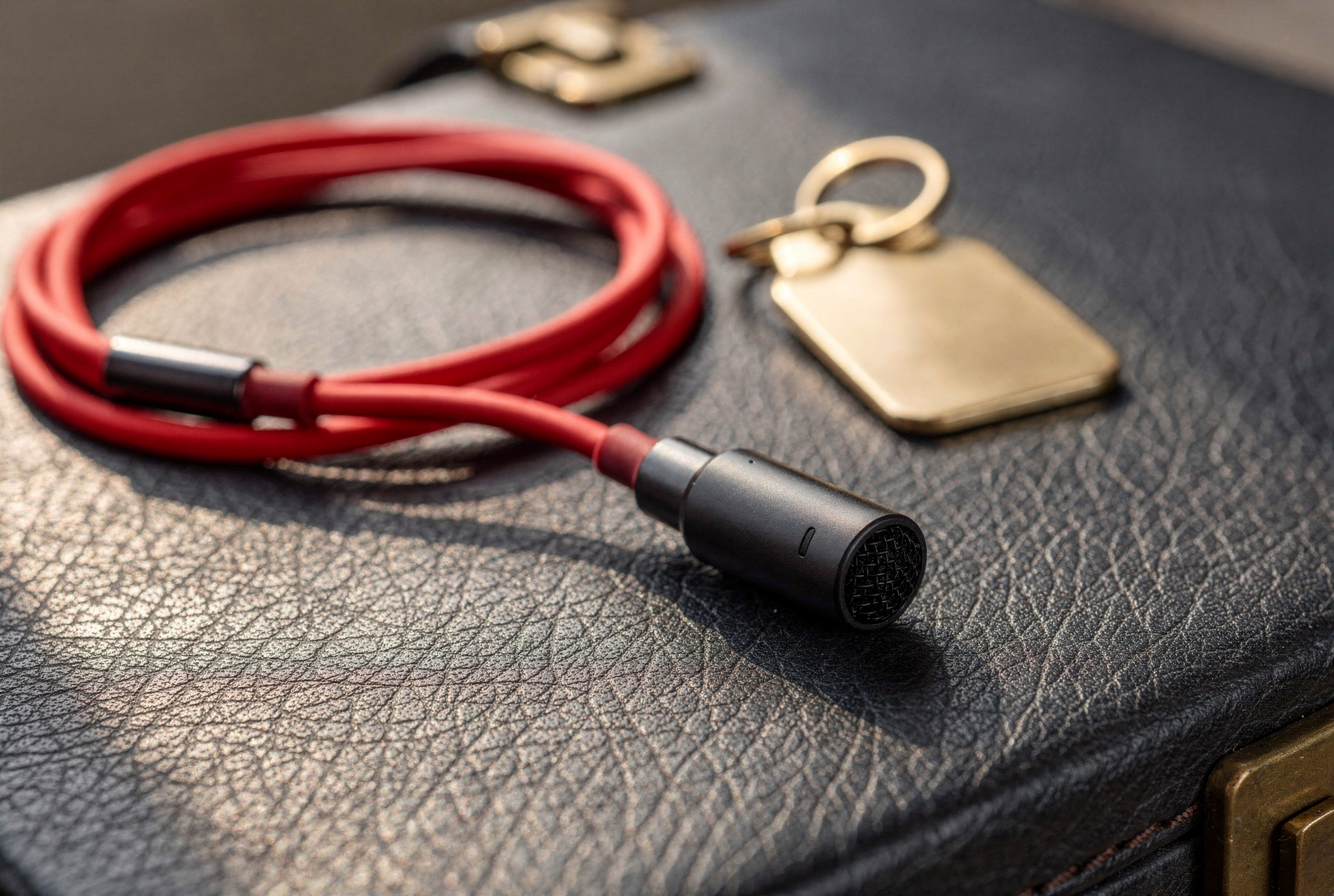 A black clip-on lavalier microphone and coiled red charging cord resting on a textured dark leather briefcase.