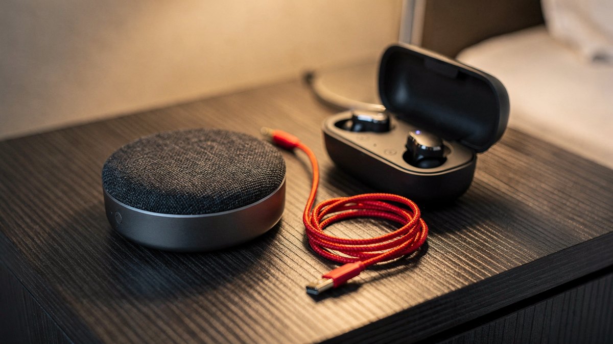 A round graphite business conference speakerphone puck and open wireless earbud case resting beside a coiled red cable on a hotel bedside console.