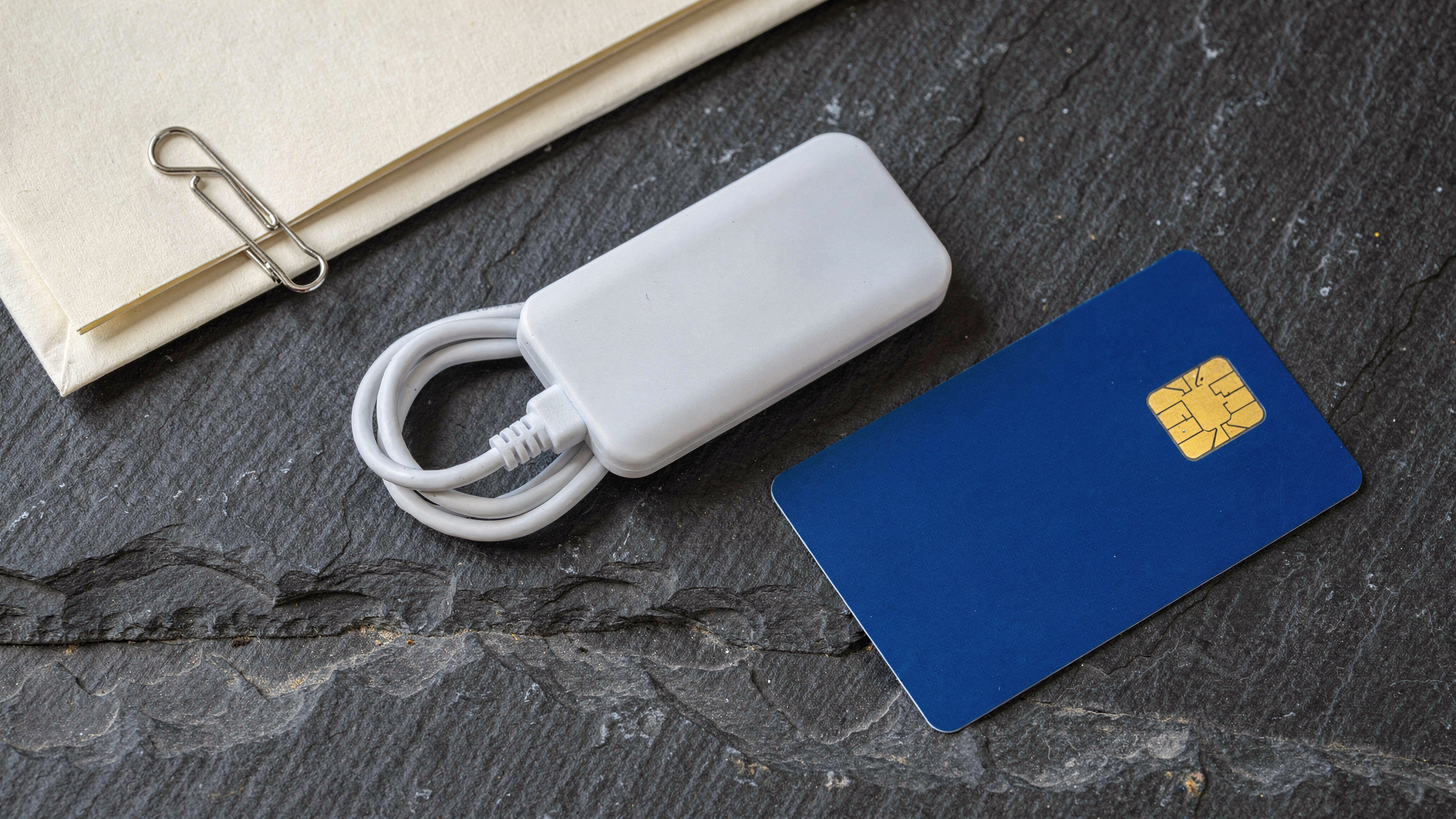A white USB smart card reader and a cobalt blue smart card with gold microchip contacts rest on a charcoal slate stone surface alongside clipped cream documents.