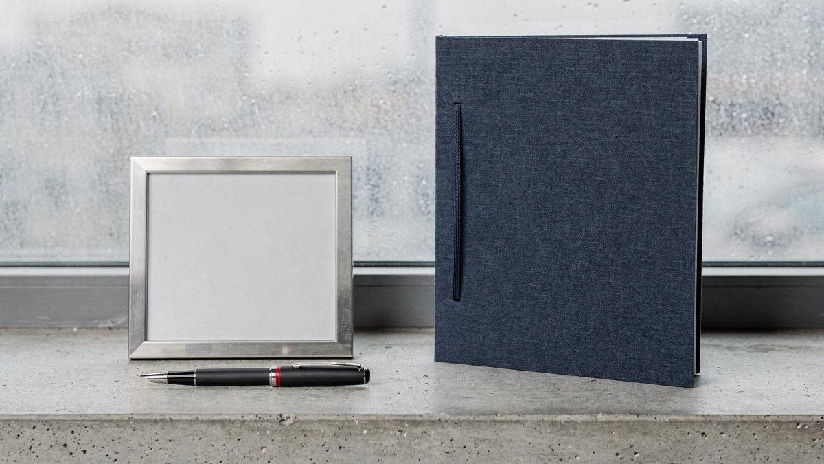 A closed dark blue dossier folder, a pen with a red accent ring, and a stainless steel photo template arranged on a concrete windowsill in flat grey overcast light.