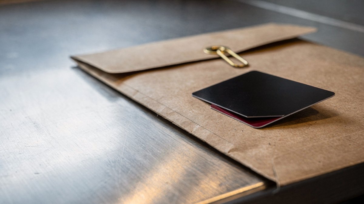 A matte black metal payment card resting on a kraft cardboard mailer over a brushed stainless steel shelf.
