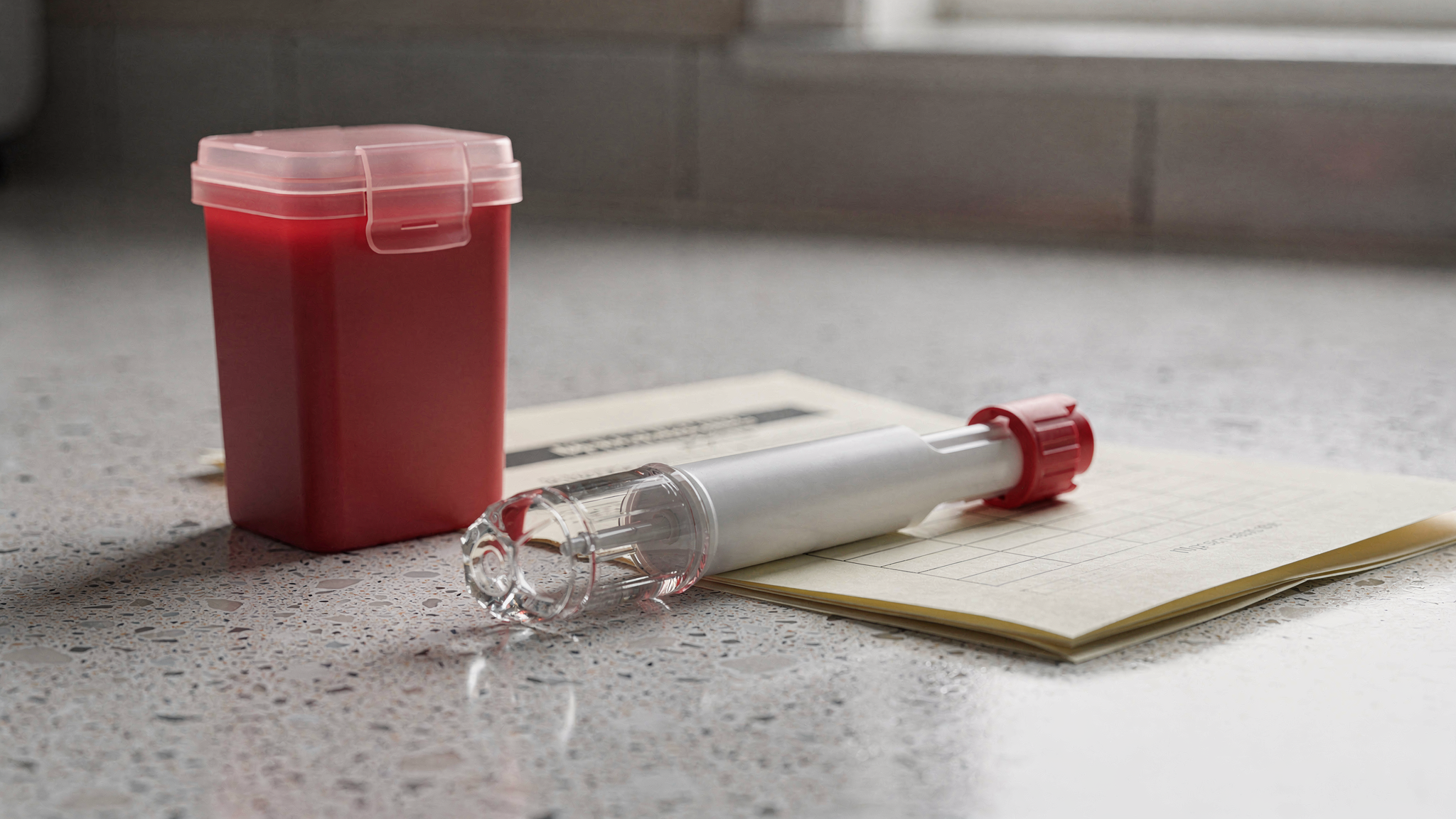 A white autoinjector pen with a red safety cap rests on a speckled countertop next to a compact red sharps container and a folded document.