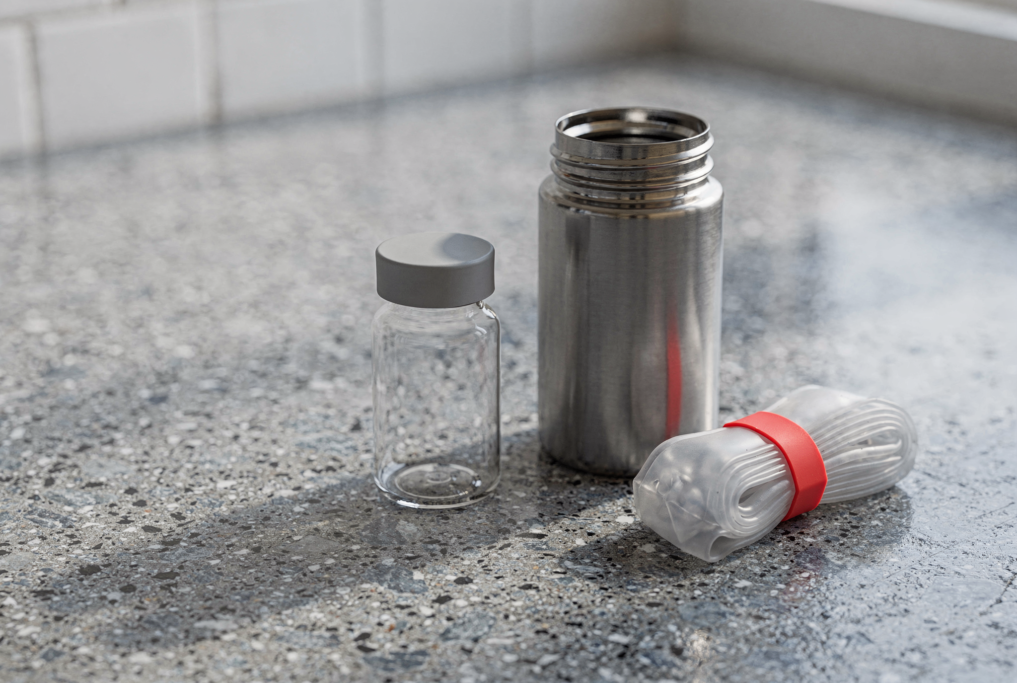 A clear glass medication vial and an open brushed stainless steel vacuum flask sit on a grey terrazzo counter alongside a cold gel pack with a red band.