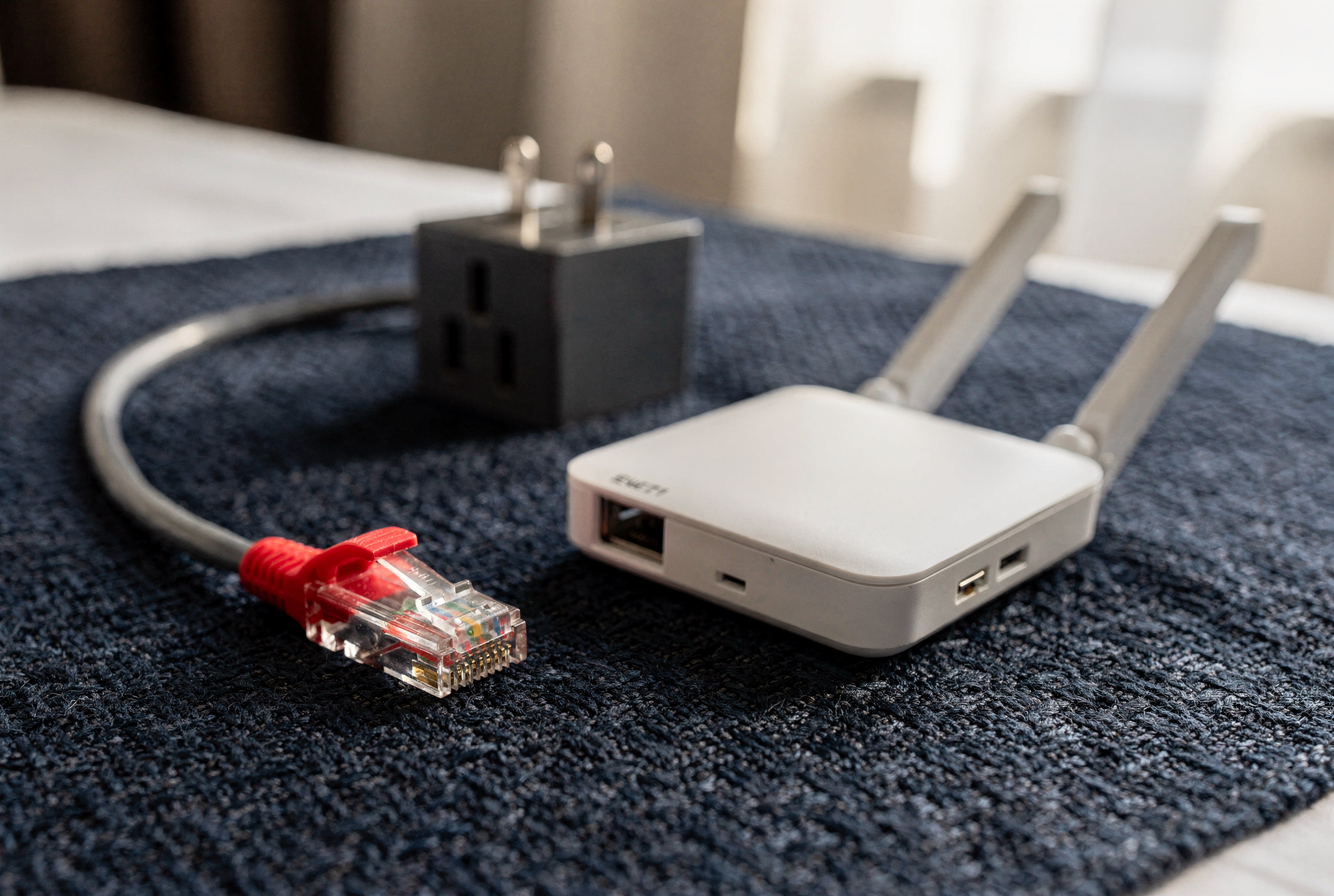 A compact portable travel router and a grey ethernet cable with a red connector boot resting on a textured navy fabric surface.