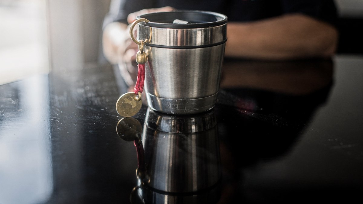 A mirrored reflection across a dark glossy cafe table showing a stainless steel travel tumbler and a key fob with a red nylon loop under soft window daylight.