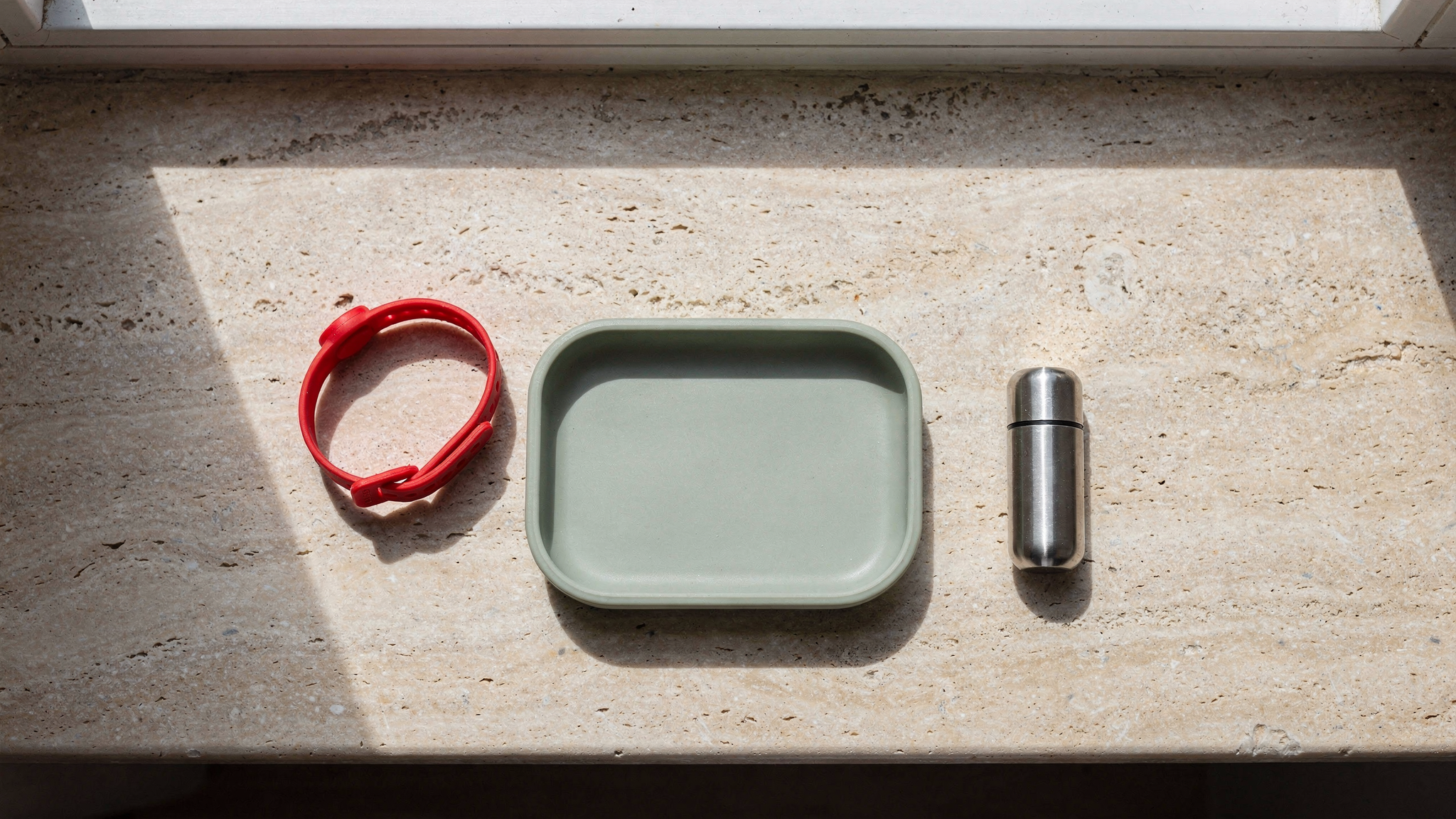 Top-down view of a sage silicone toddler plate, a red child ID wristband, and an aluminum travel pill case on a stone windowsill.