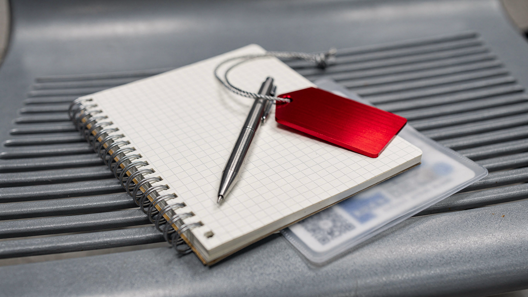 A pocket grid notebook, a stainless steel mechanical pencil, and a red aluminum luggage tag rest on a ribbed airport bench.