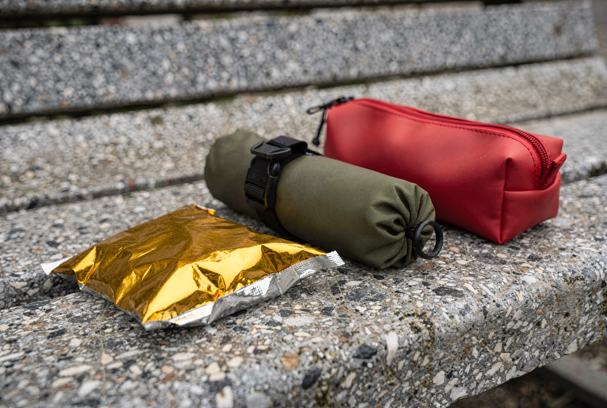 An emergency foil blanket pack, a rolled olive dry bag, and a small red silicone pouch sitting on a weathered stone bench.