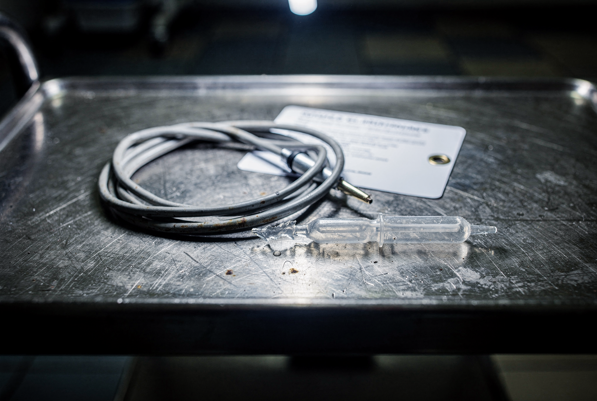 A clinical thermometer probe, open sterile saline ampoule, and laminated claim card rest on a steel tray under a harsh single night light.