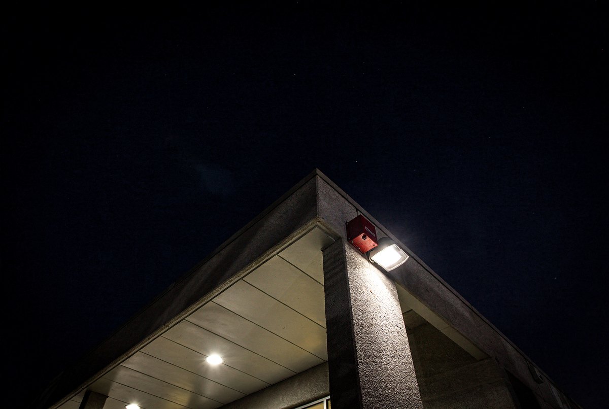 Low-angle view looking straight up at the geometric concrete awning of a night-time clinic lit by a single harsh wall lamp against the dark sky.