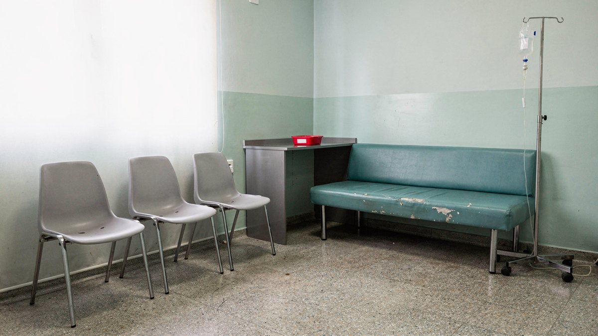 An empty outpatient medical clinic waiting room with pale chairs and an IV stand bathed in soft daylight.
