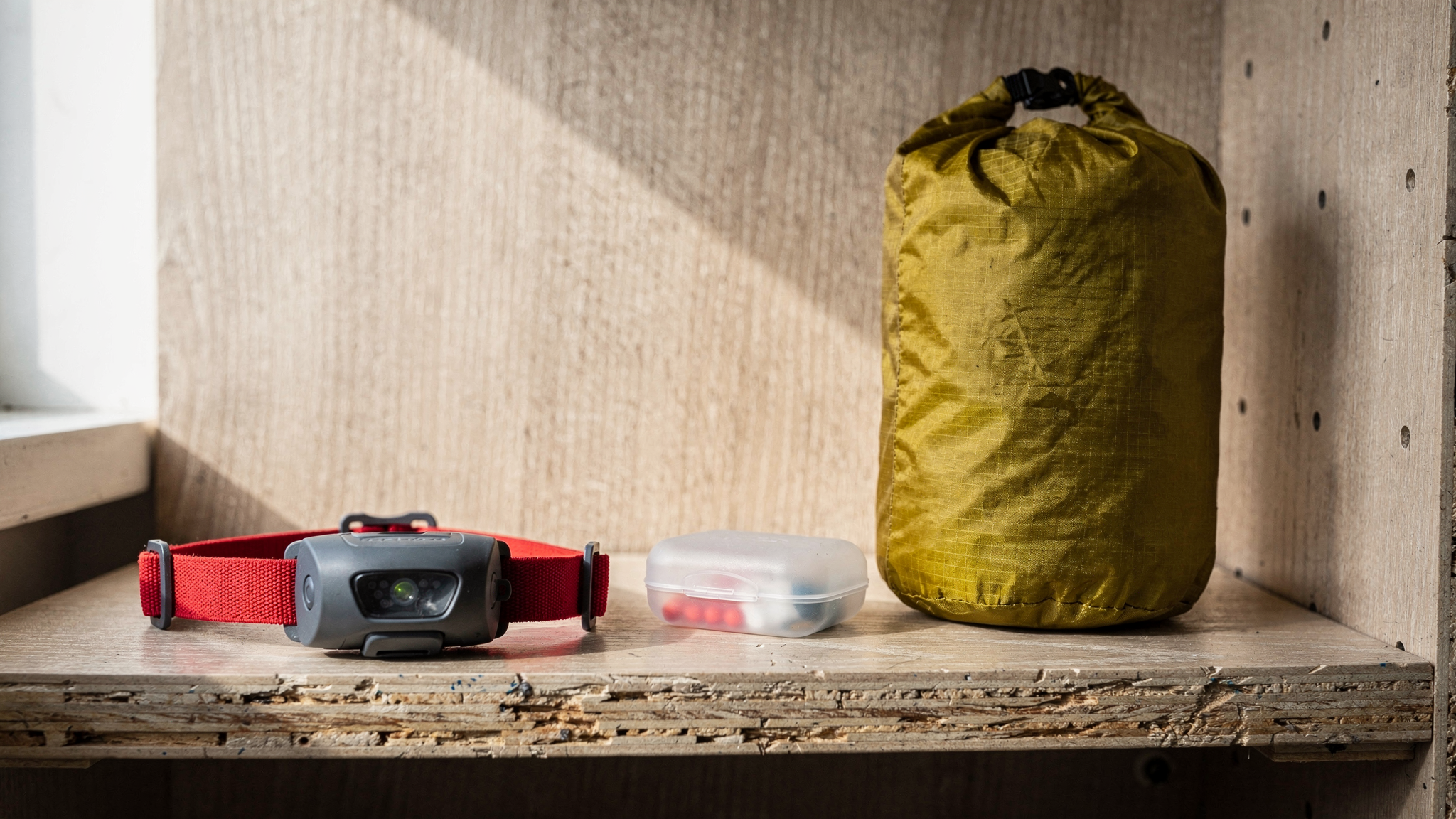 A packed ultralight dry bag and a trail headlamp with a red elastic strap resting on a wooden hostel bunk ledge.