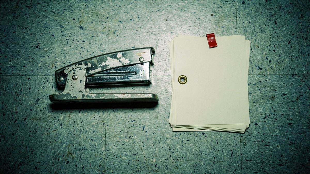 A heavy steel stapler and eyelet-bound paper dossier resting under sterile fluorescent light on a speckled linoleum surface.
