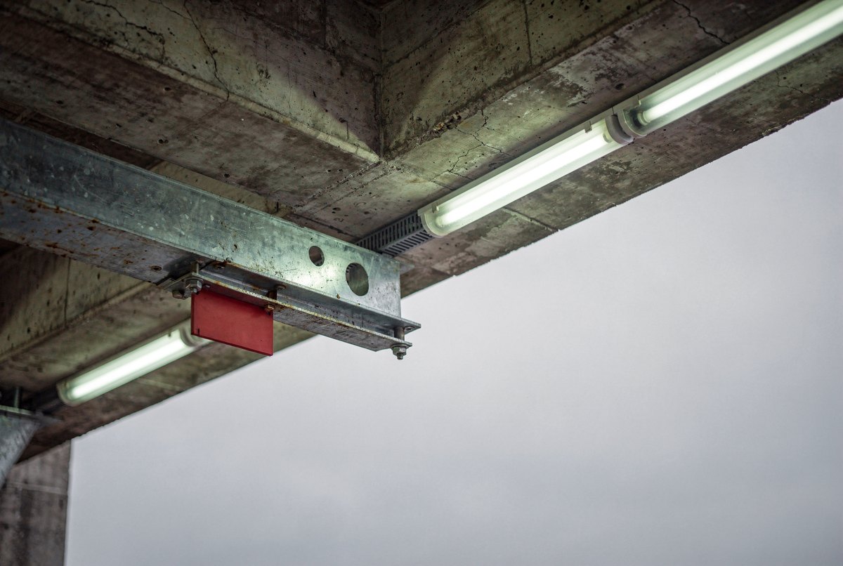 A low-angle view looking up past a concrete overhang and steel conduit bracket lit by cool fluorescent light toward an overcast sky.
