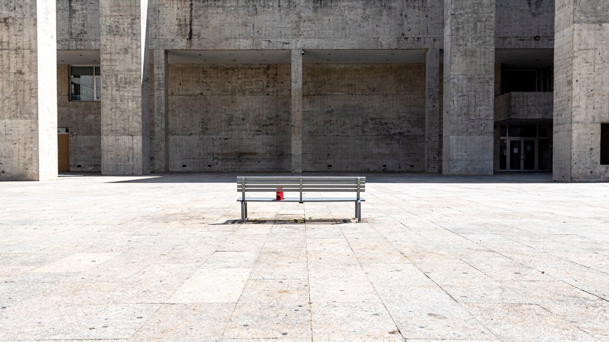 A solitary red queue clip rests on an empty public bench across a vast, sun-bleached concrete courtyard under direct overhead sun.