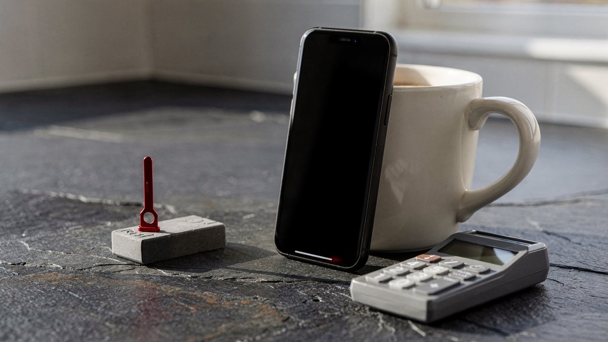A black smartphone resting against a ceramic mug next to a portable banking card reader and a red SIM pin on a slate surface.