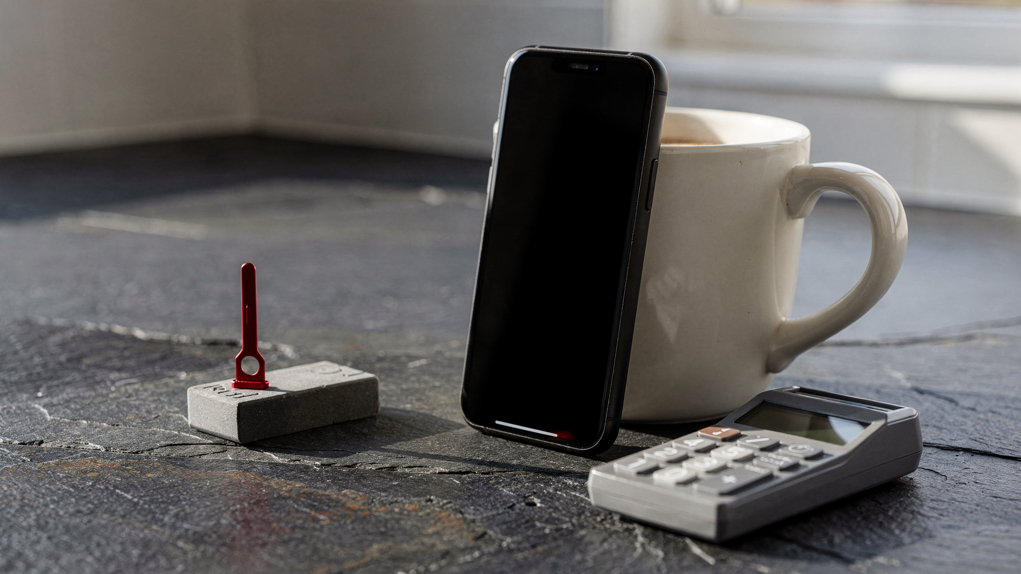 A black smartphone resting against a ceramic mug next to a portable banking card reader and a red SIM pin on a slate surface.