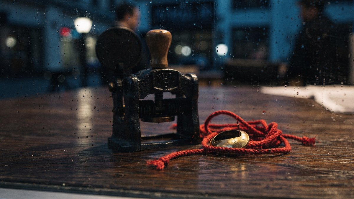 A cast-iron desk embosser and crimson cord seen through a rain-streaked office window during blue hour.